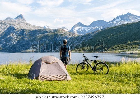 Man standing next to his trekking tent and bike on a bike travel through Norway with fjord and mountains in the background