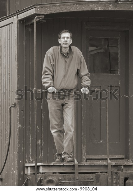 Man standing on the back of an old train car\
caboose, leaning on the\
rail.