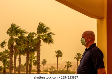 A man is standing on a balcony in the hotel and has a face mask to protect him from the Calima sandstorm, which brings a lot of fine dust. Concept: health and travel