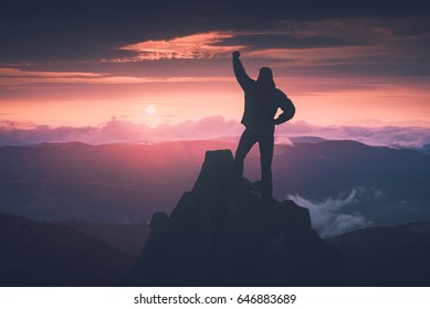 Man standing on a cliffs edge with raised hand against colorful sunset in a mountain valley. Instagram stylization. - Shutterstock ID 646883689
