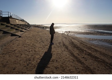 A Man Standing On Crosby Beach In Liverpool, England. He Is Standing Under The Sun And Looking Out To Sea