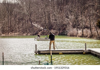 Man Standing On Dock Fishing While Another Man In A Bass Boat Floats By On Small Lake In Early Spring