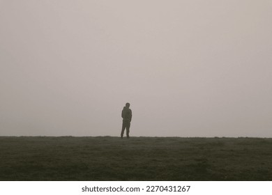 Man standing on hill in the mist and fog, a mysterious figure with fog creating a dreamy, atmospheric, mysterious scene on an outdoor field with dewy mist around a man standing alone on a hill - Powered by Shutterstock