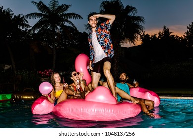 Man Standing On Inflatable Mattress And Looking Forward With Friends Sitting At Back. Group Of Men And Women Partying In The Pool At Night.