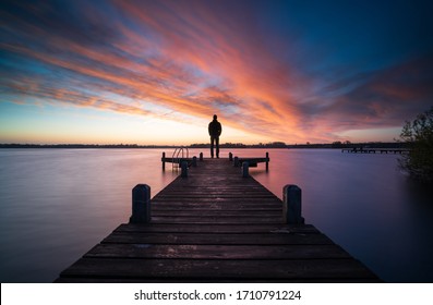 Man standing on a jetty looking over a lake during a colorful dawn.