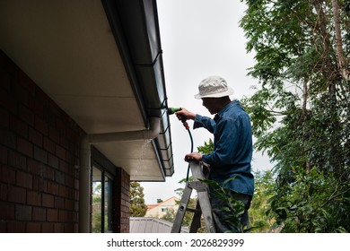 Man Standing On The Ladder And Washing The Gutter Using A Garden Hose. Home Maintenance Work. 