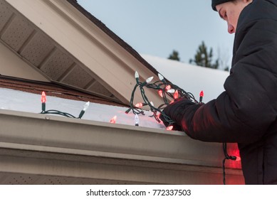 Man standing on ladder while hanging red and white Christmas lights on roof of house.