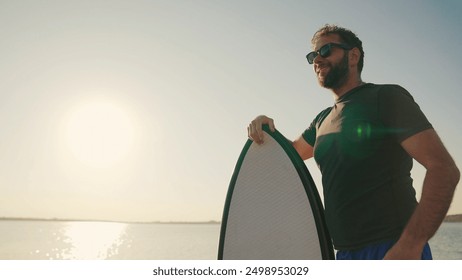 A man standing on a lake pier at sunset, leaning on a water board. Male surfer holding a short surfboard while standing on a pier with a motorboat in the background - Powered by Shutterstock