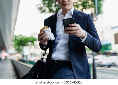 Man standing on the outdoor street. Eat burger while sending e-mail and playing social network with smart mobile phone.