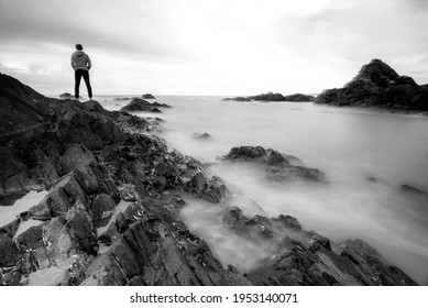 Man Standing On A Rocky Coastline, A Slow Shutter Speed Was Used To See The Movement . Soft And Grain Image.