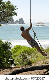 A Man Standing On A Rope Swing And Admiring The Beautiful Sea View During His Summer Vacation.