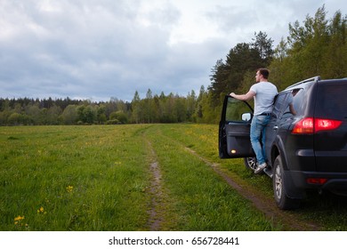 The man standing on the running Board of the car with the door open and looks into the distance. Traveler stands on step of all-terrain 4x4 vehicle before board barn.