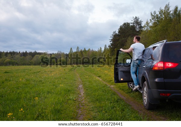 The man standing on the\
running Board of the car with the door open and looks into the\
distance. Traveler stands on step of all-terrain 4x4 vehicle before\
board barn.