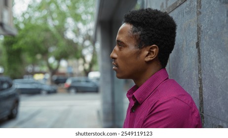 Man standing on urban street with stone wall background and cars in the distance, wearing checkered shirt, captured mid-day in city environment with green trees visible. - Powered by Shutterstock