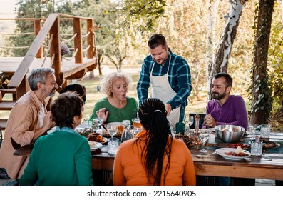 Man Standing Over The Dining Table With Friends And Family,  Serving A Pot With Delicious Food  Group Of Diverse People For Holiday Festive Dinner Party On The Outdoor Kitchen
