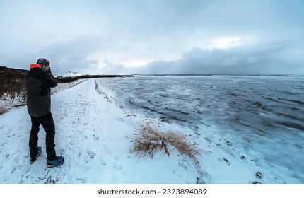 Man standing protected by the polar cold with jacket, hat and boots, taking photos of a completely snow covered road and landscape and a frozen river in Iceland - Powered by Shutterstock