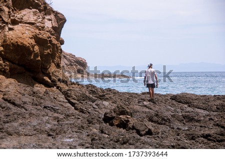 A man standing at the rocks looking at the horizon at the sea surrounded by rocks. Blue ocean. Blue mountains far away. Socialdistancing.