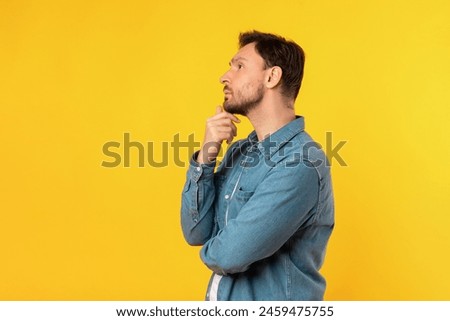 A man standing upright in front of a solid yellow background. The man is facing forward with a neutral expression, touching his chin and looking at copy space