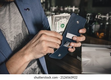 A Man Stands Against The Background Of The Cafe's Interior And Takes Dollar Bills Out Of His Wallet To Pay For A Business Lunch, Close-up. Horizontal Orientation, No Face, Copy Space