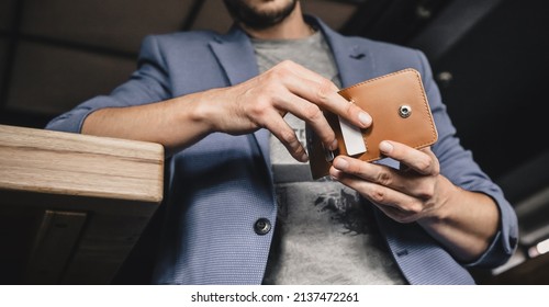 A Man Stands Against The Background Of The Interior Of The Cafe And Takes Out A Plastic Card From His Wallet To Pay For A Business Lunch, Close-up. Horizontal Orientation, No Face, Copy Space