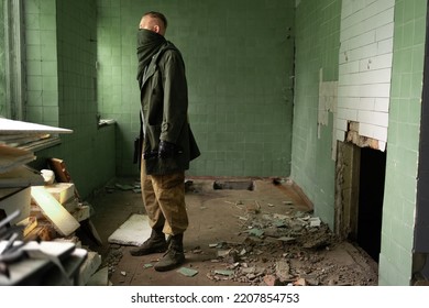 Man Stands Among Ruins Of A Destroyed Building, Holding A Gun And A Flashlight In Hands. Concept Of Survivor After End Of World Or Third World War Is Looking For Shelter And Food Supplies.