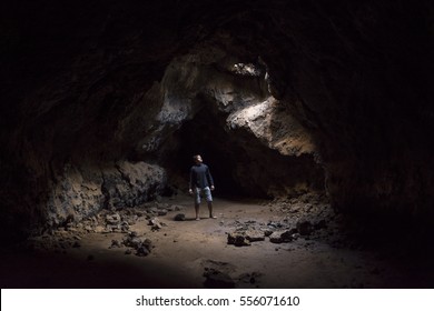 Man stands in eerie underground cave looking up at hole in ceiling pouring in sunlight at Mojave lava tubes in California - Powered by Shutterstock