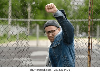 A man stands in front of a chain-link fence, raising his fist in a gesture of solidarity. He wears a denim jacket and glasses, exhibiting a determined expression in daylight - Powered by Shutterstock