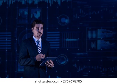 A man stands in front of a large screen displaying data visuals. He holds a tablet in one hand and gestures as he speaks. The setting is a conference or meeting room with low light.