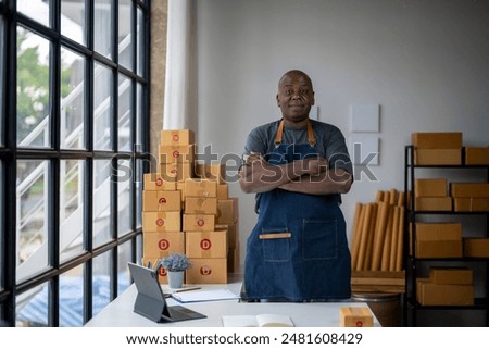 A man stands in front of a stack of boxes with a blue apron on. He is wearing a black shirt and has his arms crossed. The scene appears to be a small business office or warehouse