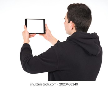 Man Stands With His Back And Holds A Tablet With Empty Screen. Young Man Demonstrates A Tablet Computer With Space For Text On The Screen. Guy In Black Clothes Holds An Electronic Tablet At Eye Level.