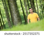 A man stands in a lush, green forest, wearing a yellow shirt blending into the serene natural environment. The tall trees around him form a peaceful, quiet atmosphere, with dappled sunlight 