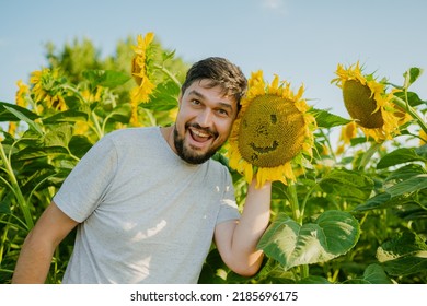 A man stands near a sunflower and smiles, rejoices at a good harvest. A young farmer stands in a sunflower field and checks the harvest. Agricultural concept