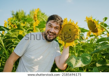 A man stands near a sunflower and smiles, rejoices at a good harvest. A young farmer stands in a sunflower field and checks the harvest. Agricultural concept
