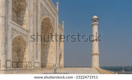 A man stands next to the Taj Mahal. The hand is raised, looking up. The scale of the structure is visible. White marble mausoleum and minaret against the blue sky. Agra. India. 