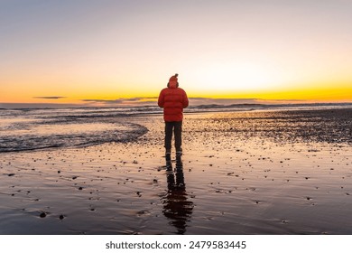 A man stands on a beach at sunset, wearing a red jacket. The scene is serene and peaceful, with the man's reflection in the water adding to the calm atmosphere - Powered by Shutterstock