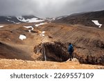 A man stands on the edge of a cliff against the background of a waterfall, mountains and a volcano, red-yellow volcanic sand, Kamchatka.