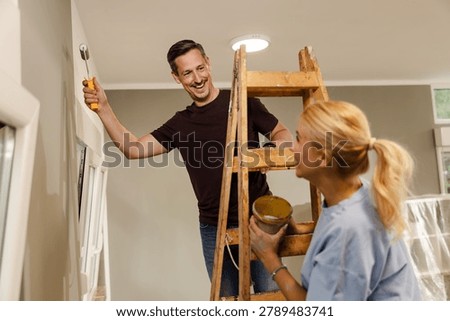 A man stands on a ladder while painting the ceiling. A woman holds a paint can and smiles at him. They are working together on their home during daytime hours.