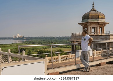 A man stands on the observation deck of the Red Fort next to the jasmine tower Musamman Burj. In the distance, among the green fields, the white marble Taj Mahal is visible. Reflection in the river 