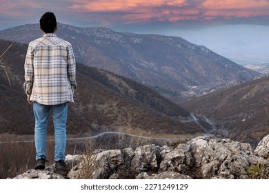 A man stands on a rock overlooking a valley and looks out at the sunset.