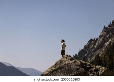 A man stands on a rocky mountain top, looking out at the horizon. The sky is clear and blue, and the mountains in the distance are majestic. The man is lost in thought - Powered by Shutterstock