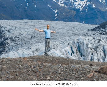 A man stands on a rocky outcropping, arms outstretched, overlooking a massive glacier with deep crevasses and mountains in the background in Iceland. - Powered by Shutterstock