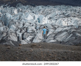 A man stands on a rocky outcropping, dwarfed by the expansive white and blue ice formations of a glacier in Iceland. The scene captures nature's beauty and power. - Powered by Shutterstock