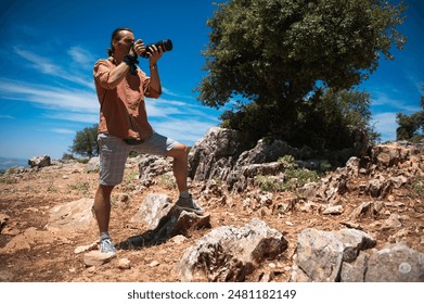 A man stands on a rocky terrain, capturing photos with a DSLR camera. The bright, sunny sky and rugged landscape create a scenic backdrop. - Powered by Shutterstock