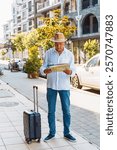 man stands on a sidewalk holding a map and a suitcase, observing the surroundings in a vcity.