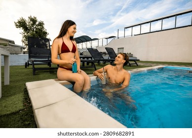 A man stands in the swimming pool while his partner sits on the edge, talking and laughing together with two cocktail bottles placed next to them.