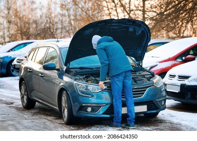 Man Stands Under Open Hood Of Modern Car, Checking Engine Oil Level In Winter Season On Parking Lot. Man Check Fluid Levels In Winter. Winter Car Care