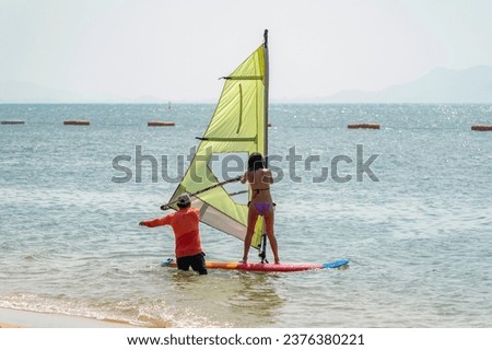A man stands in the water and teaches a woman to ride a sailboard at a tropical resort. A woman stands on a red board and holds a yellow sail.