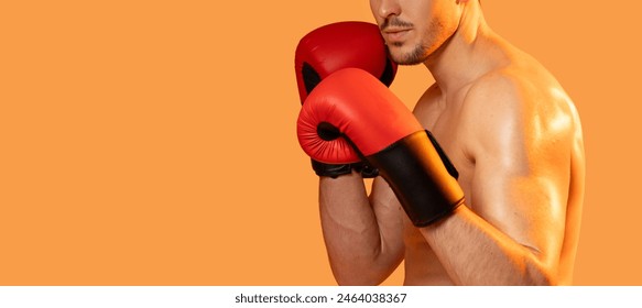 A man stands wearing boxing gloves against a vibrant orange background. He appears ready for a fight, with determination in his stance, cropped, panorama with copy space - Powered by Shutterstock