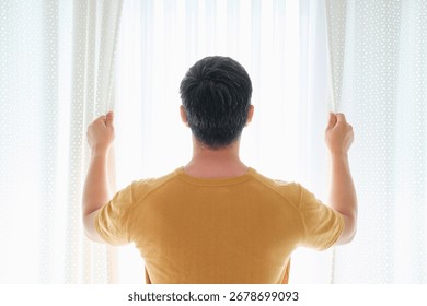 Man stands at window and opens white curtains, rear view.