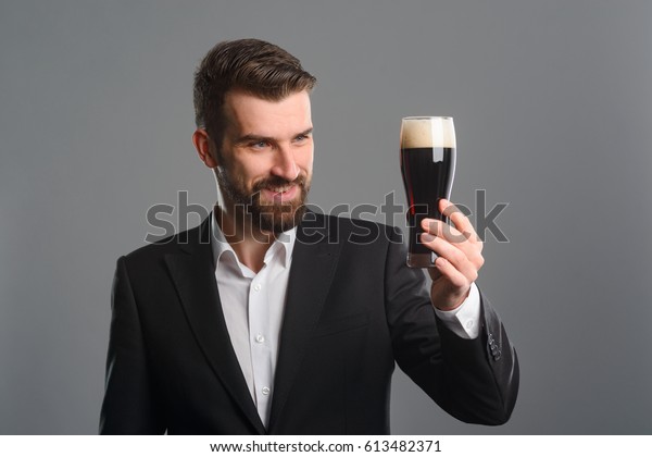 Man\
staring at beer glass. The beer is excellent: dark brown color of\
liquid and thick foam of head. Studio shot of satisfied bearded guy\
in office style clothes holding a glass of\
beer.